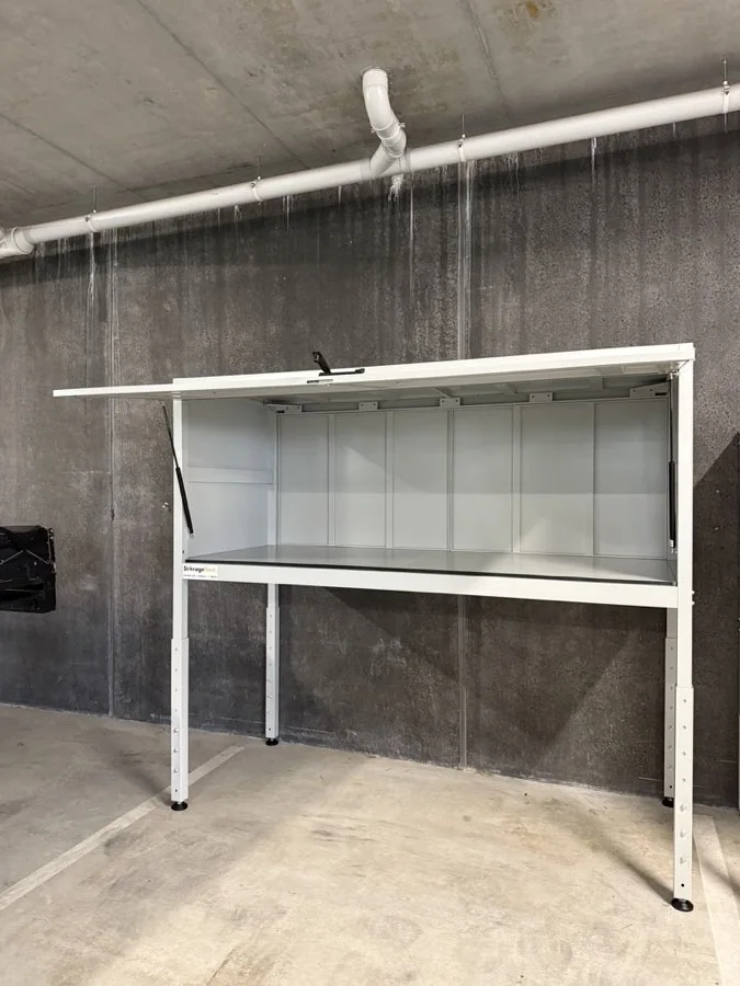 A Kookaburra over bonnet storage unit standing open against a concrete wall, showing the full width of its steel storage compartment